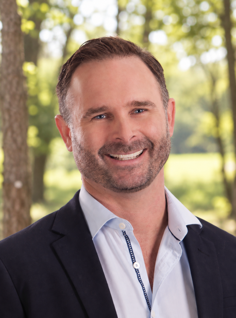 Head-and-shoulders portrait of a smiling man in a navy blazer and light blue shirt, outdoors with green trees in the background.