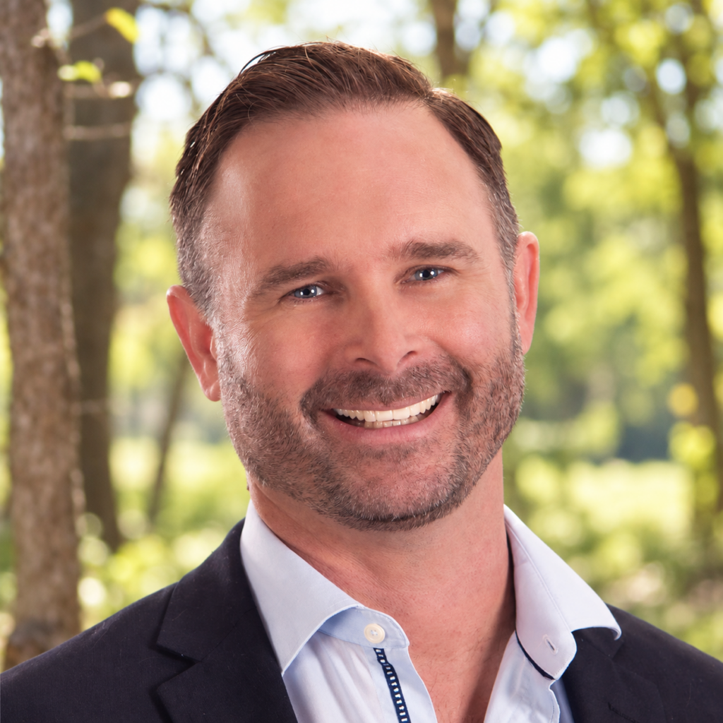 Head-and-shoulders portrait of a smiling man in a navy blazer outdoors with green trees in the background, sunny day