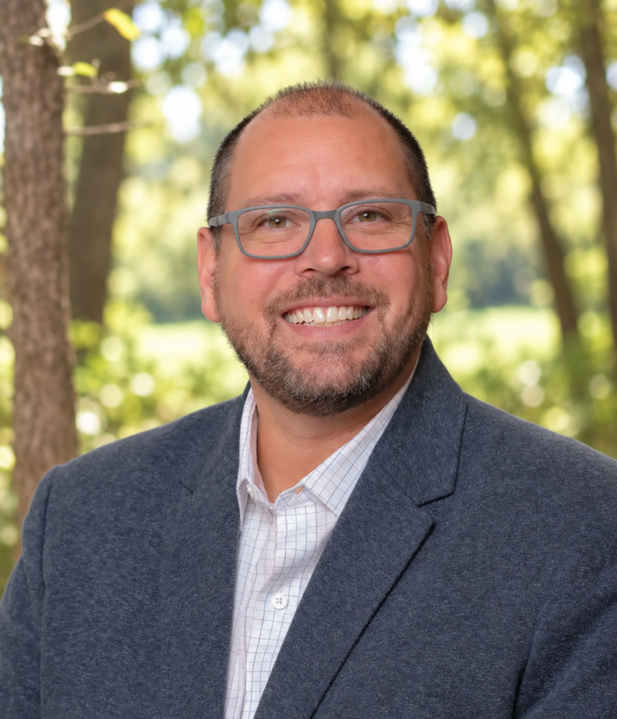 Smiling man in a blue blazer and gray glasses posing outdoors in a sunlit park setting.